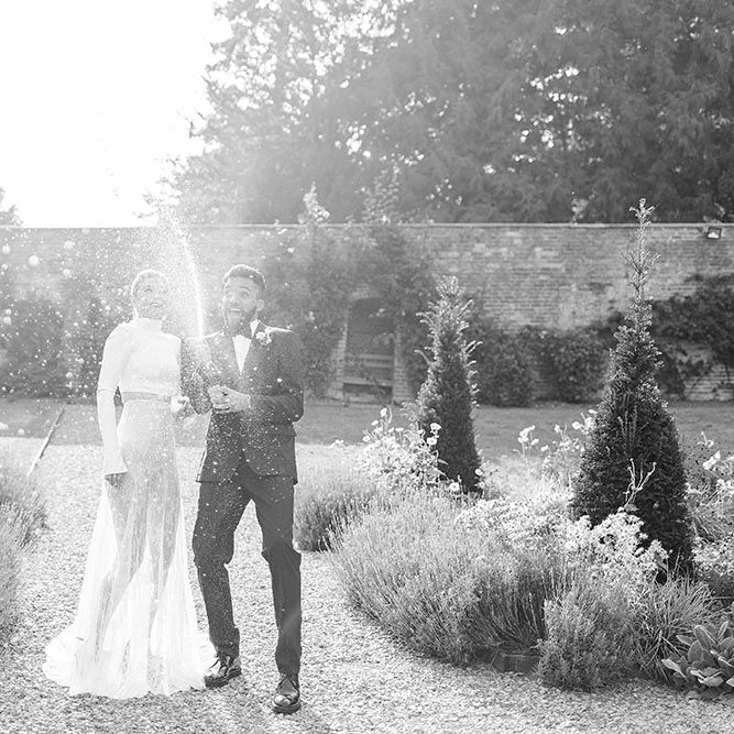 Bride in separates and groom in a tuxedo spraying champagne in the gardens at Garthmyl Hall