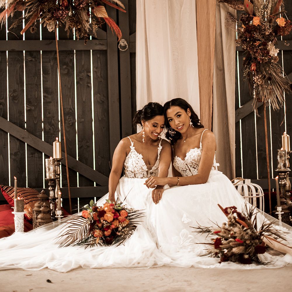 Two brides in tulle skirt dress with lace bodices sitting on the floor at The Barn at Drovers with dried and fresh flower installations and candle and lantern floor decor 