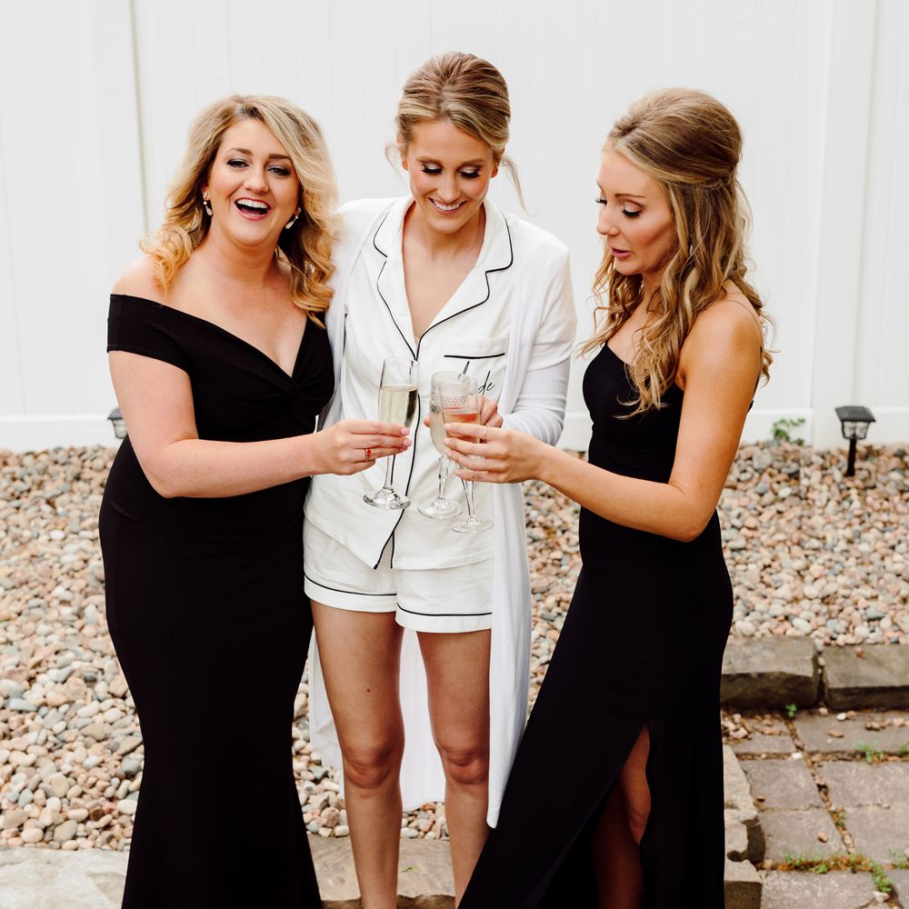 Bride in short white satin pyjamas and white fluffy slippers stands outside with two bridesmaids in black bridesmaids dresses all clinking champagne flutes before backyard wedding