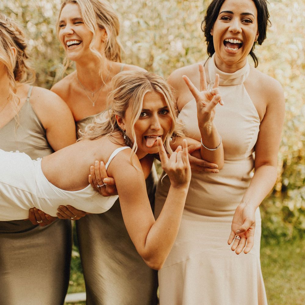 Bridesmaids in sage green and beige bridesmaids dresses hold bride in white Made With Love wedding dress as she sticks out tongue during wedding reception at Anran Devon