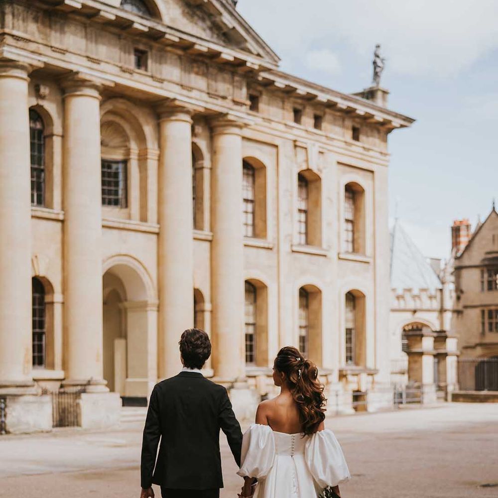 Stylish bride with a full ponytail wearing a strapless wedding dress with full skirt and button back detail holding hands with her groom in a tuxedo 