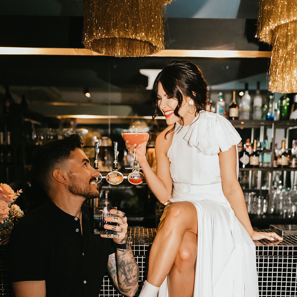 City wedding with groom in a black short sleeve shirt and bride in a stylish wedding dress with boots sitting on the bar drinking cocktails 