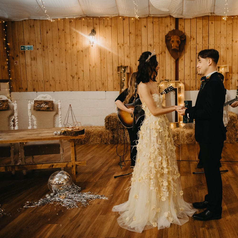 Bride in strapless feathered wedding dress and headpiece holds hands with groom in dark blue suit during rustic wedding ceremony