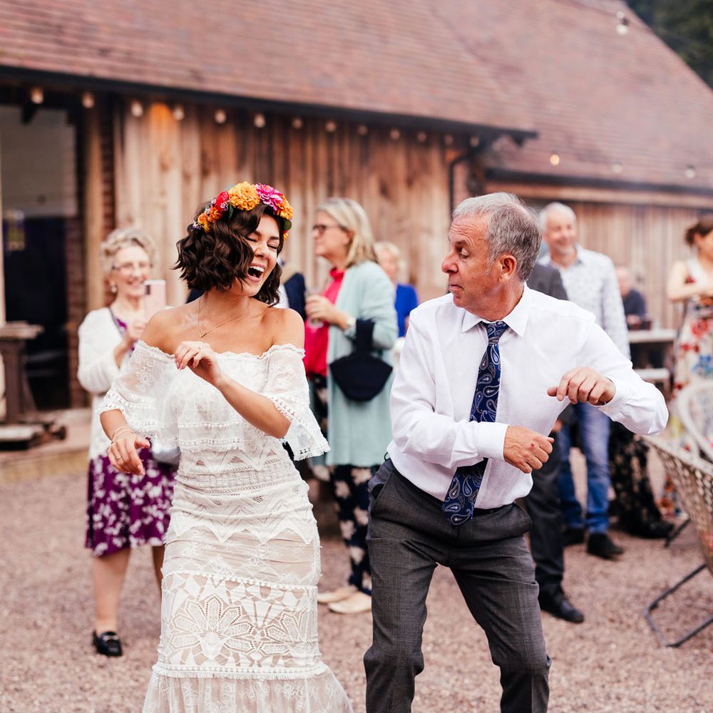 Bride in strapless Grace Loves Lace wedding dress and colourful flower crown dances with guests in grey trousers, white shirt and blue paisley print tie outside at rustic barn wedding