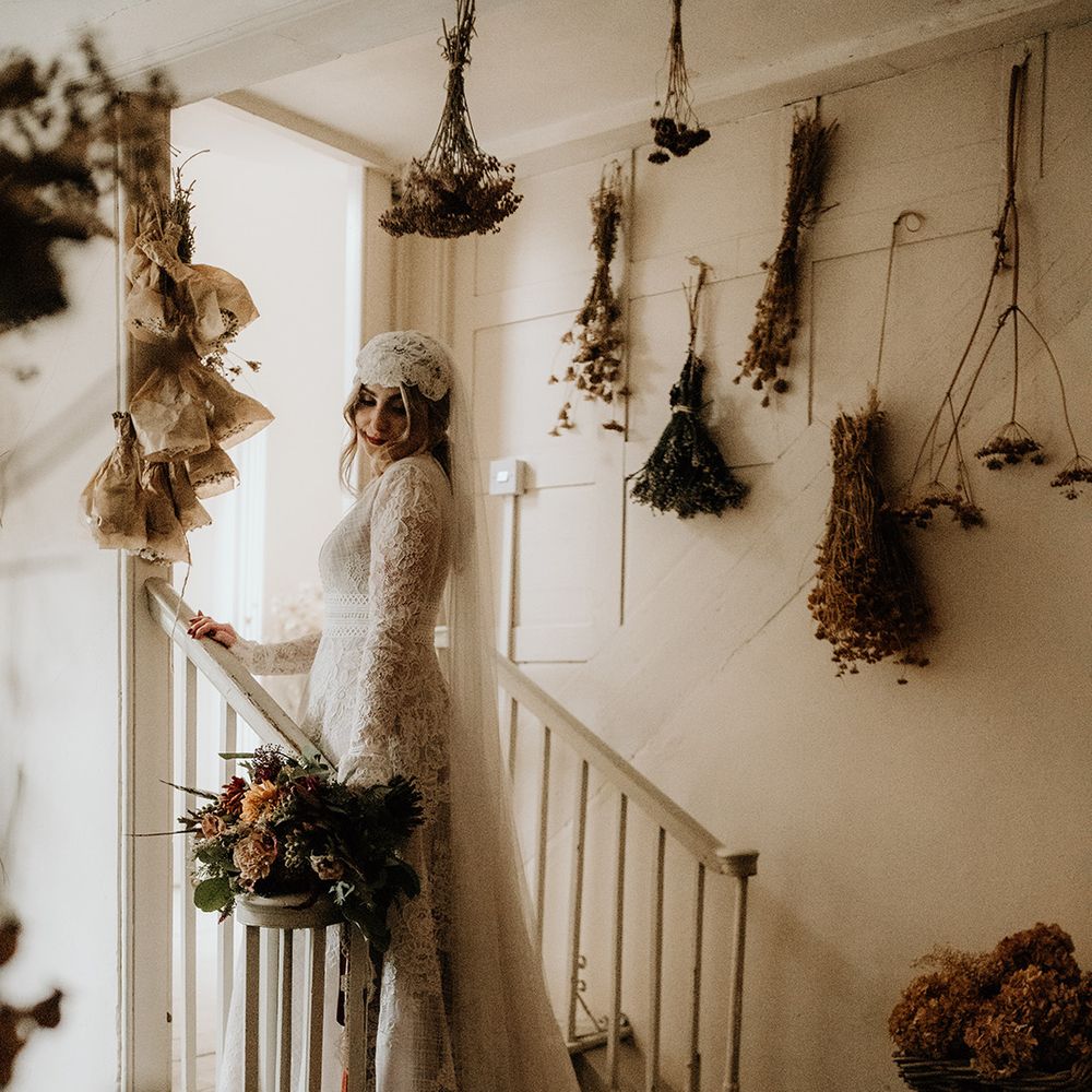 Bride in a lace wedding dress with plunging neckline and bell sleeves at Lyde Court wedding with hanging dried crops on the wall 