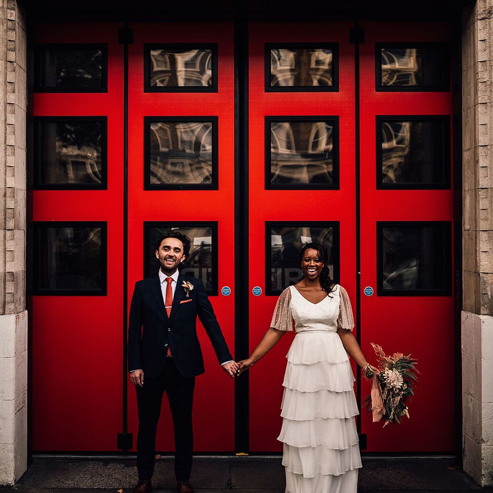 Black bride in a layered skirt and pearl top holding hands with her groom in a navy suit in front of Brixton station's red doors 