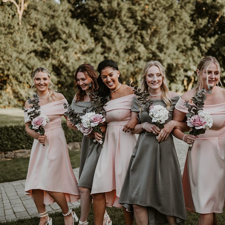Bridesmaids wear off the shoulder tea length bridesmaids gowns in sage green and pastel pink whilst holding floral bouquets