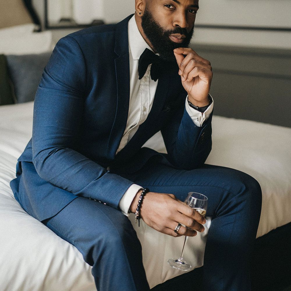 Black bearded groom in a navy blue suit with black bow tie sitting on the bed on the wedding morning 
