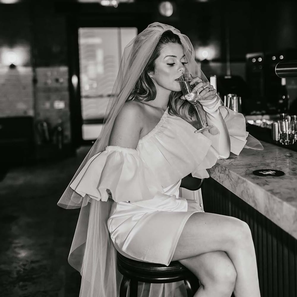 Bride in a ruffle off the shoulder short wedding dress sitting at the bar drinking Champagne 