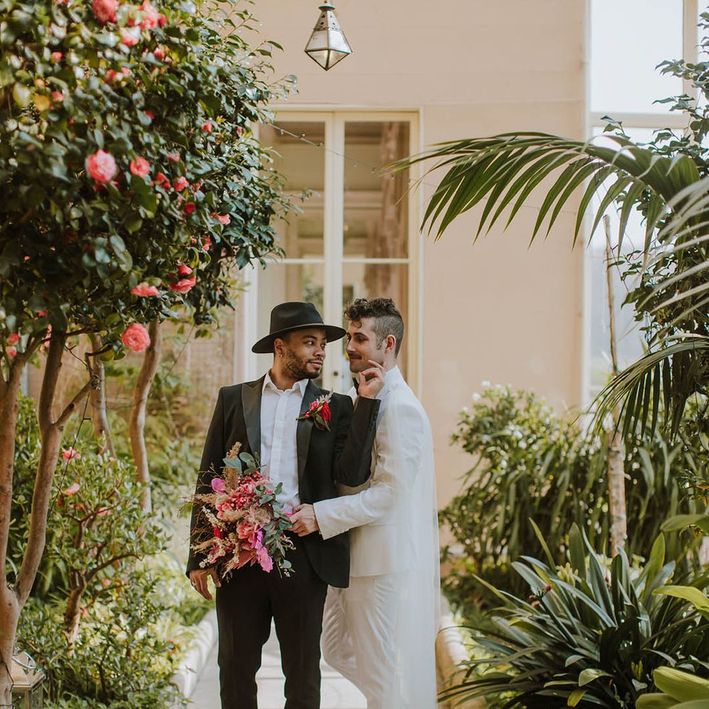 Groom in a black suit and fedora hat standing with his partner in a white groom suit and cape holding a vibrant red bouquet 