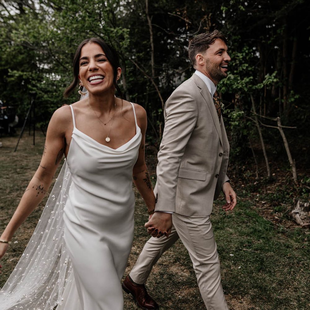 Groom in a stone coloured suit holding hands with his bride in a satin slip wedding dress and pearl veil 