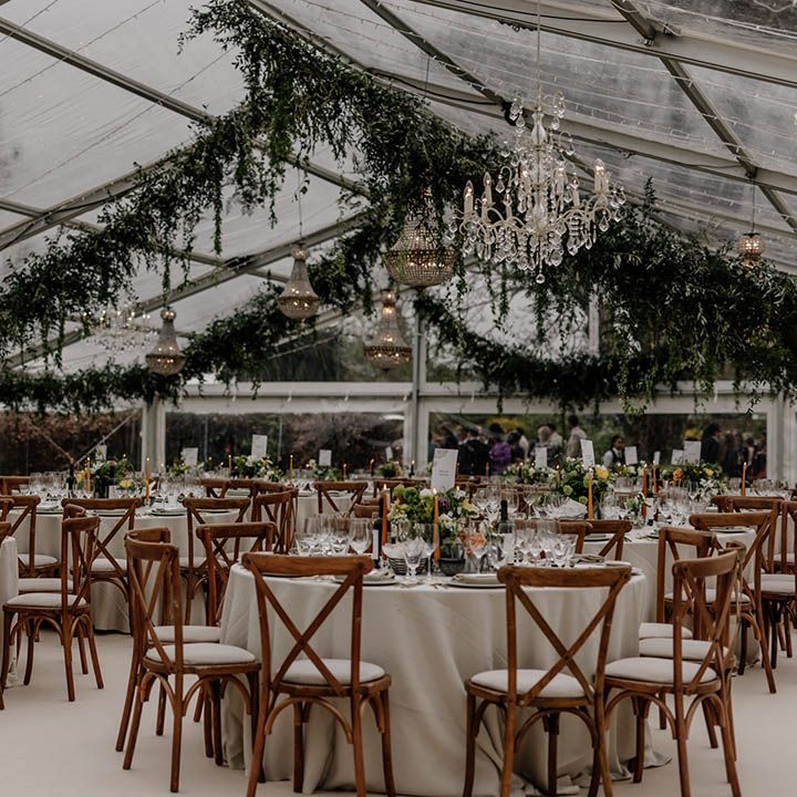 Clear glass marquee wedding reception with wooden furniture, greenery decor and handing chandelier 