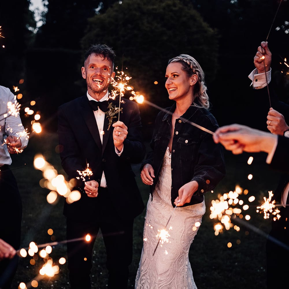 Bride and groom wave sparklers with their guests with the bride wearing a black denim cover up 