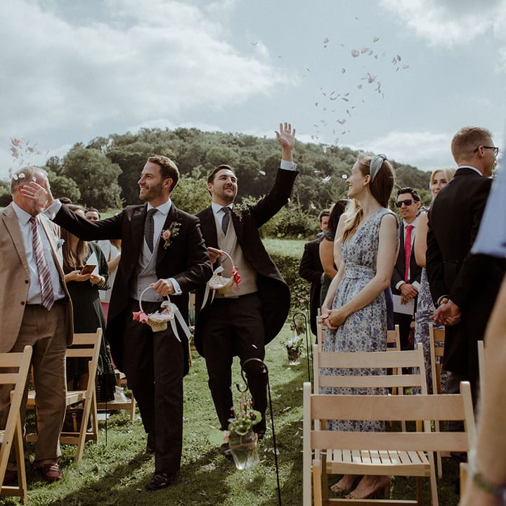 Groomsmen in morning suits walk down the aisle with wicker baskets with pink ribbons throwing out petals 