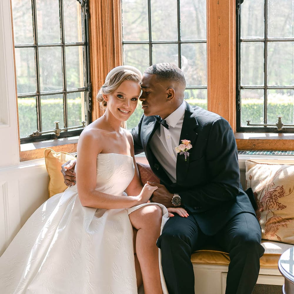 Groom sits on the window seat with the bride and rests his forehead against her as she smiles at the camera 