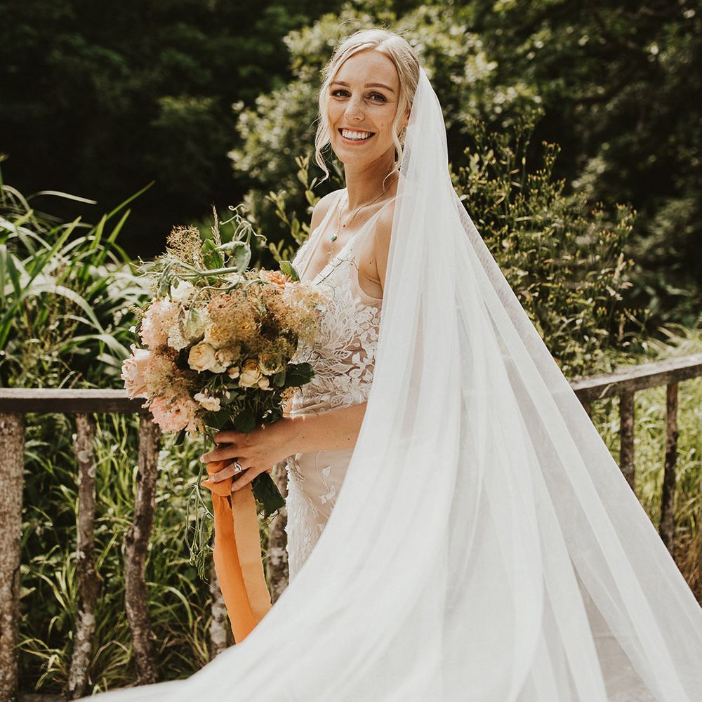 Bride with a long veil in a lace wedding dress wearing a blue pendant necklace holding an orange coloured bouquet 