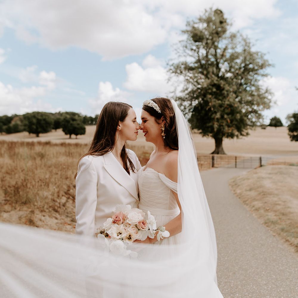 Bride in white suit smiling at the bride in an off the shoulder wedding dress holding a pale pink and white rose and orchid bouquet 