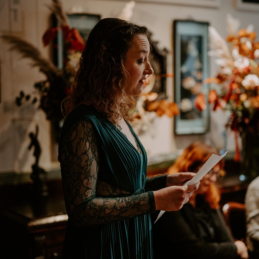 Bridesmaid in bottle green bridesmaid dress with long lace sleeves doing reading at autumnal wedding