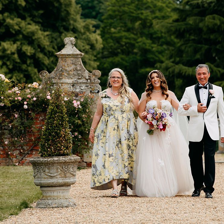 Bride in strapless tulle wedding dress and sheer bridal cape being walked down the aisle by her mother and father 