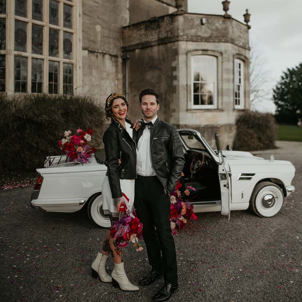 Groom in white shirt, black bowtie, black suit trousers and slim fitting black leather jacket standing by off white classic convertible wedding car embracing bride in blazer style wrap around short wedding dress with delicate fringe detailing and personalised, embroidered black leather jacket holding hot pink, bright pink and viva magenta bridal bouquet with dahlias, orchids, roses, amaryllis, anthuriums and dried flowers 
