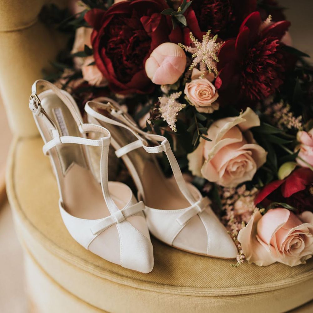 White wedding heels with a bow and an ankle strap next to the classic bridal bouquet of pink and red wedding flowers 