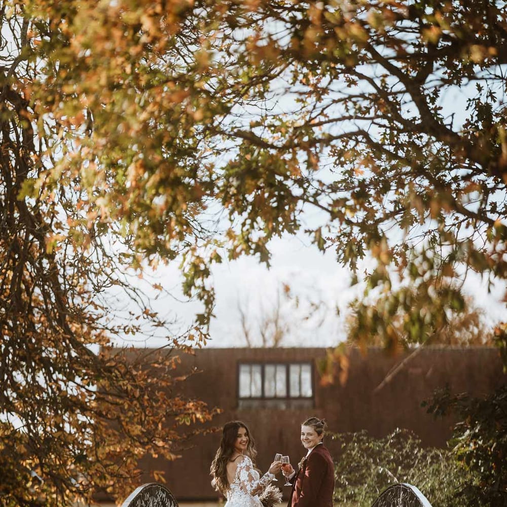 Bride in maroon bridal suit with grey waistcoat on bridge at Bassmead Manor Barns wedding venue with bride in long sleeve lace illusion sleeve wedding dress with overlay 