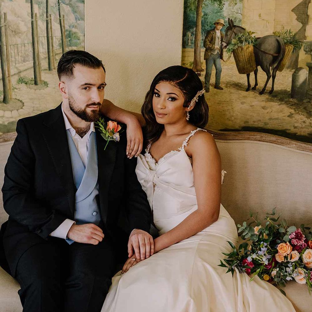 Groom in black blazer with pink garden rose and foliage boutonniere, cornflower blue waistcoat and bride in bow front sleeveless satin wedding dress with large gold bridal hair clip 
