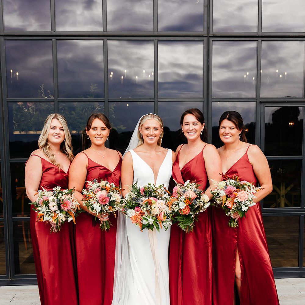 Bridesmaids in matching red satin bridesmaid dresses with leg slit posing with the bride with her curled blonde hair in sleek hairstyle with fitted wedding dress 