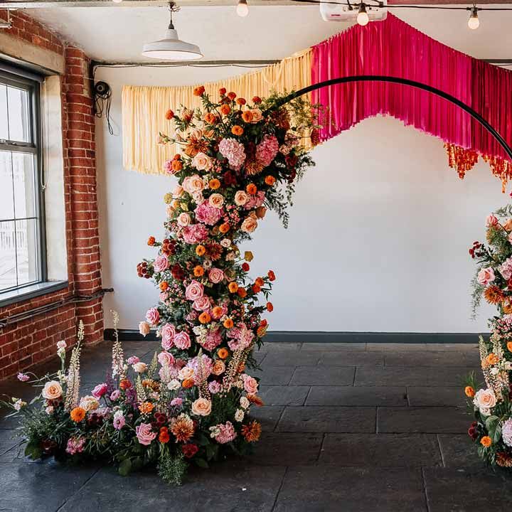 Large circular floral arch with vibrant flowers and foliage attached and pink, red, yellow and metallic wedding streamers behind at reception room of Northern Monk Refectory Leeds 