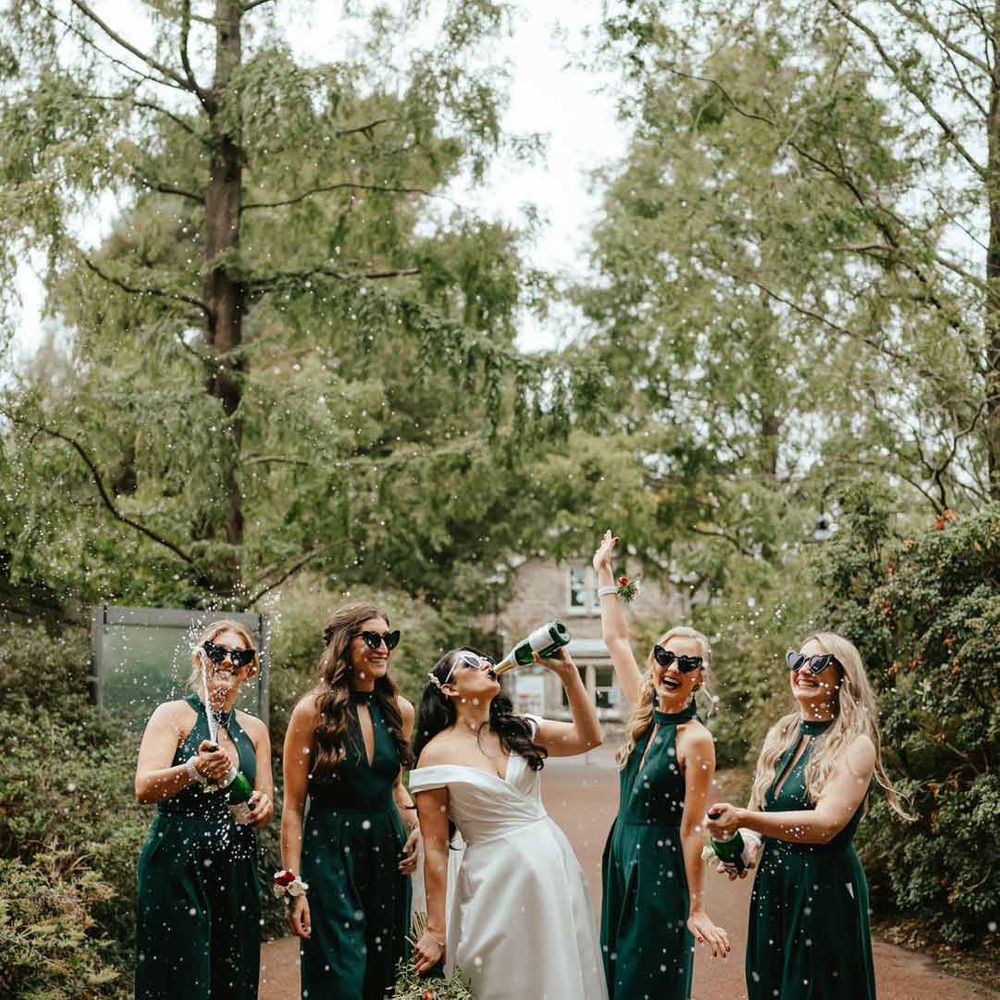 Bride in off shoulder satin wedding dress drinking champagne with bridesmaids in bottle green keyhole bridesmaid dresses with heart shaped sunglasses