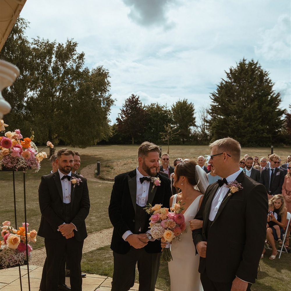 Father of the bride walks the bride to the end of the aisle with the groom and best man standing in black tuxedo with pink and orange rose floral decorations 