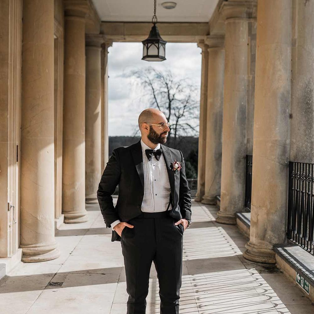Groom in classic black tuxedo with bowtie and wildflower boutonniere standing on grounds of Buxted Park wedding venue