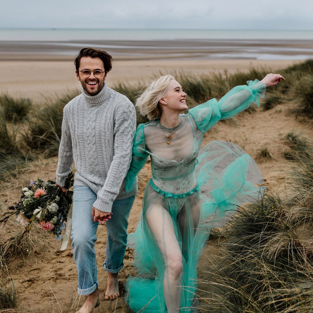 White brunette bearded groom in glasses, grey jumper and faded jeans walking through the sand dunes with his blonde bride in a sheer green pastel wedding dress and body suit