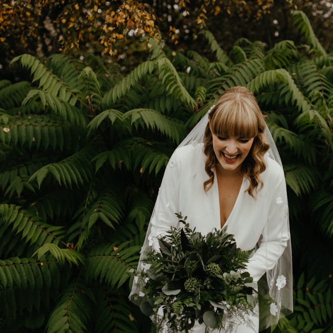 Smiley bride in a jumpsuit and appliqué veil holding a green foliage wedding bouquet