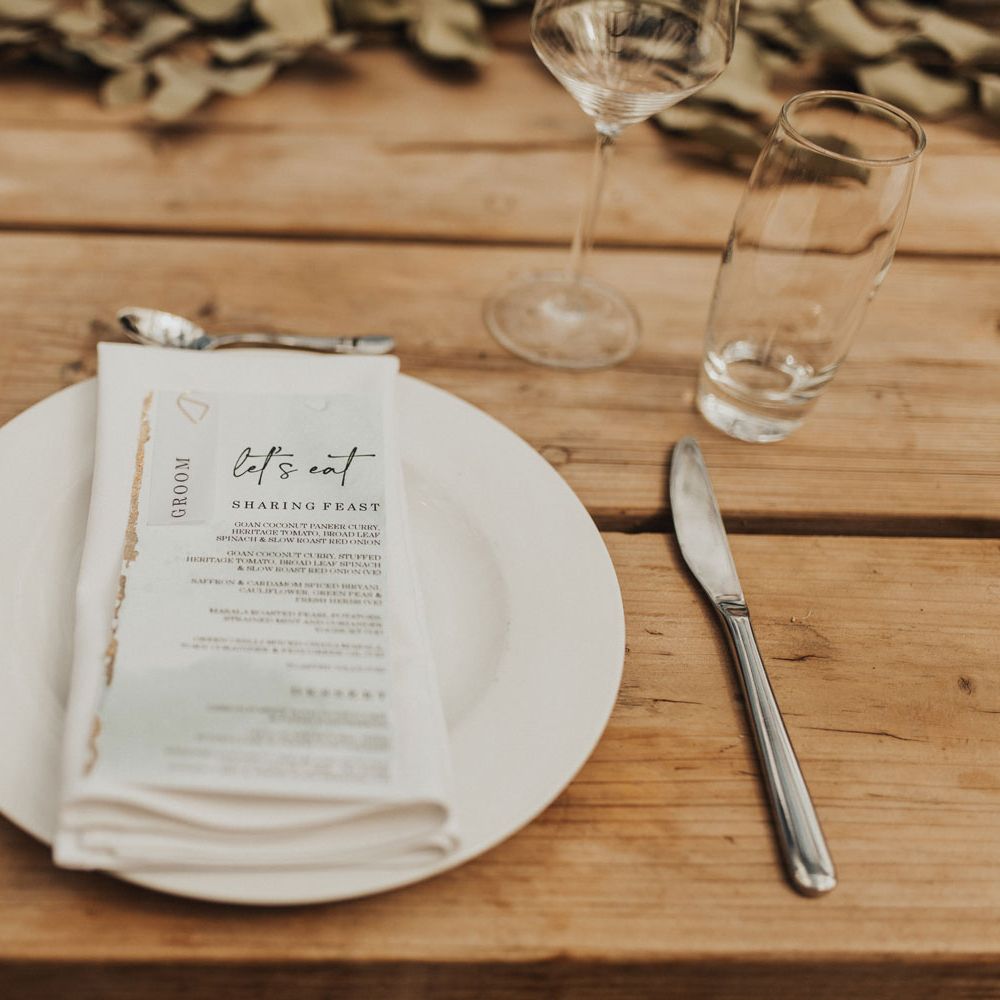 White plate setting with napkin and menu on wooden table set with cutlery and glassware with foliage table garland for wedding breakfast at Anran Devon