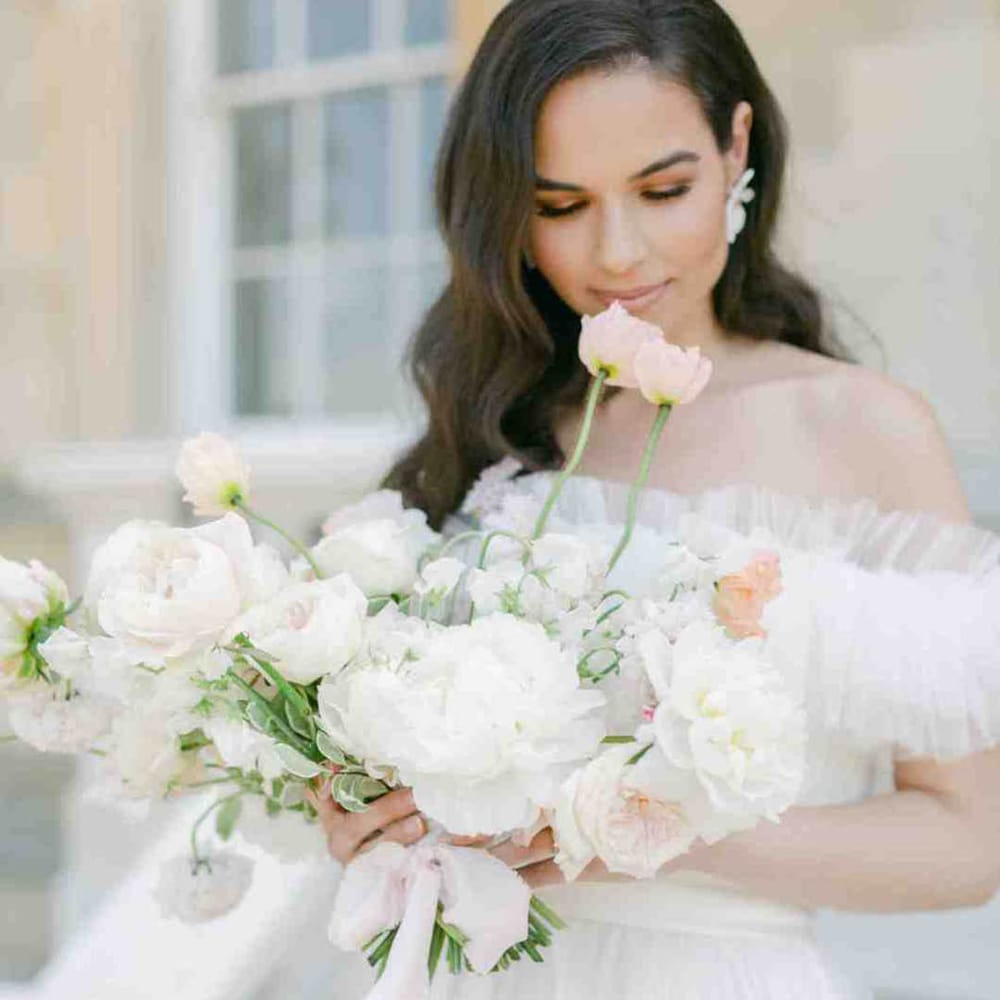 Bride with long wavy hair in an off the shoulder wedding dress looking at her pastel wedding bouquet with roses and peonies 