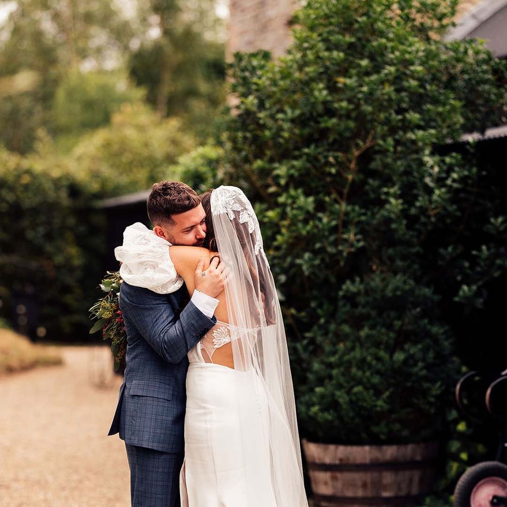 Groom in a blue check suit embracing his bride in a bespoke wedding dress with cut out back, lace puff sleeves and matching veil 