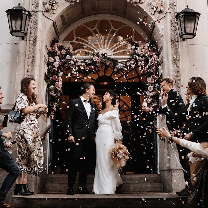 Beautiful London architectural building with blush pink flower arch with Bride and Groom cheering with wedding guests throwing colour confetti 