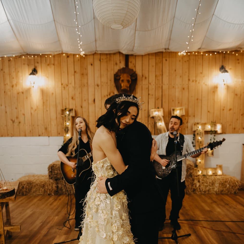 Bride in strapless feathered wedding dress and headpiece hugs groom in dark blue suit as two guitarists sing in the background during rustic wedding ceremony
