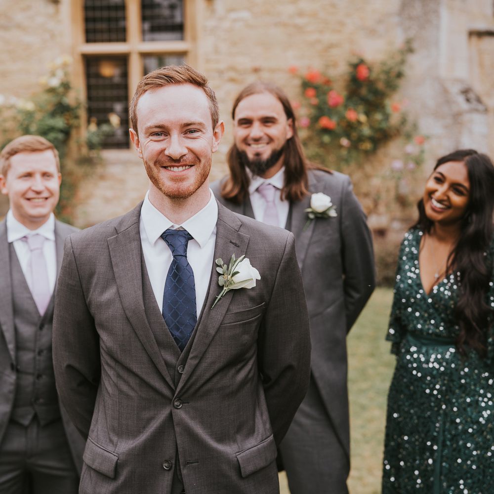 Smiling groom in grey three piece suit with blue tie and white rose buttonhole stands with groomsmen in grey morning suits and woman in emerald green sequin dress for outdoor wedding in Buckinghamshire