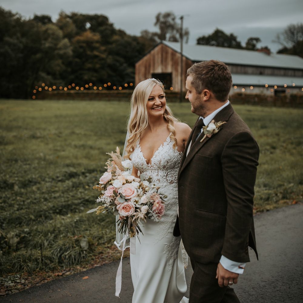 Bride in lace wedding dress and veil holding white, pink and green bridal bouquet smiles as she walks with groom in brown suit outside at Lake District wedding