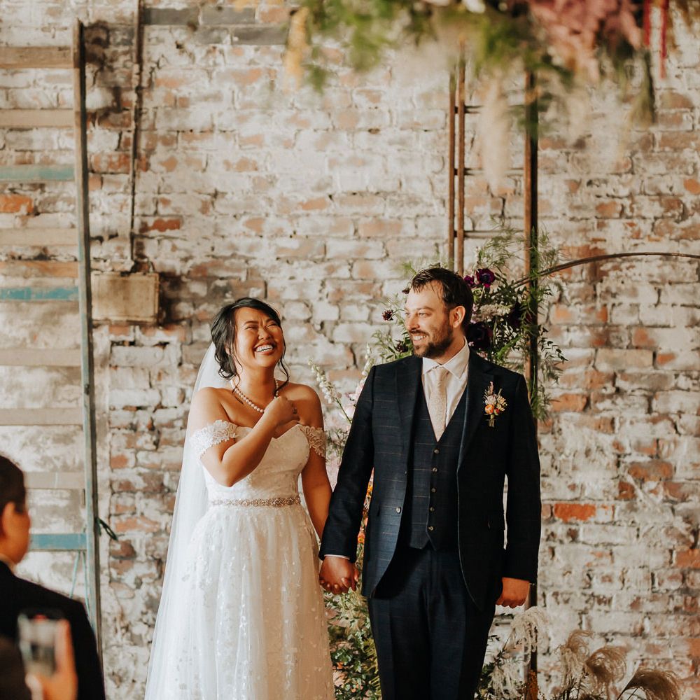 Bride in off the shoulder wedding dress and pearl necklace holds hands with groom in blue check three piece suits and floral buttonhole during wedding ceremony