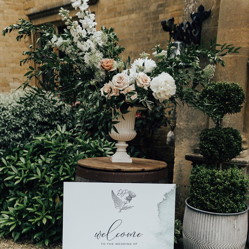 Wedding welcome sign with green watercolour design propped up against a urn floral arrangement with roses 