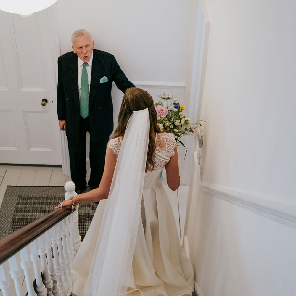 Bride walks down stairs to greet her family member in sage green tie and black pinstriped suit