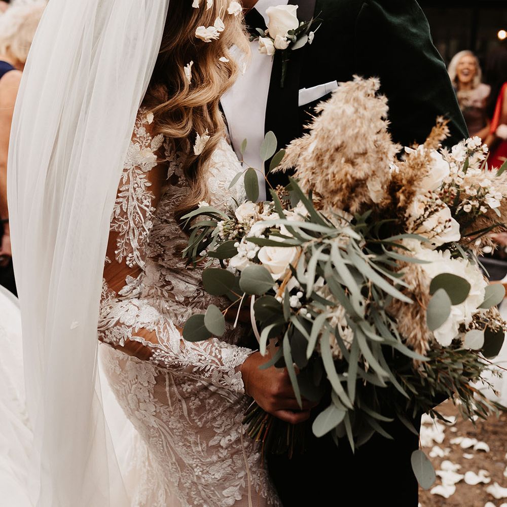 Groom in green velvet suit jacket shares a kiss with the bride who holds white rose and dried flower wedding bouquet