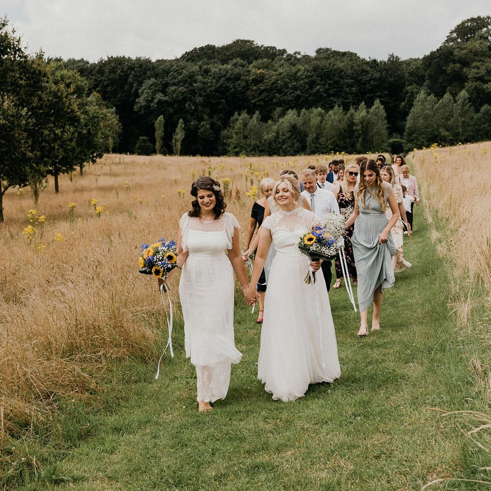 Two brides in sheer high neck wedding dresses hold hands and walk through a field as they lead their wedding guests to the barn venue