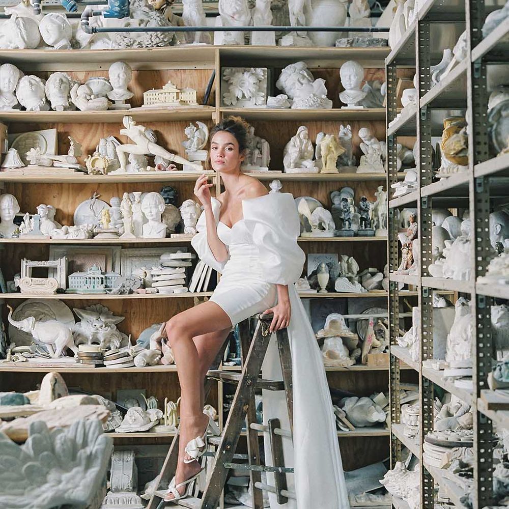 Bride seated on ladder next to figures of heads wearing short wedding dress and mule wedding shoes 