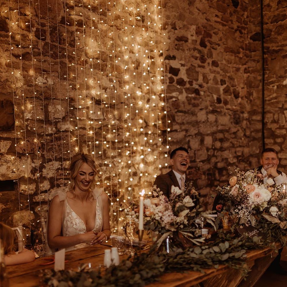 Bride and groom laughing on the wooden top table decorated with foliage runner with white candles and fairy light backdrop 