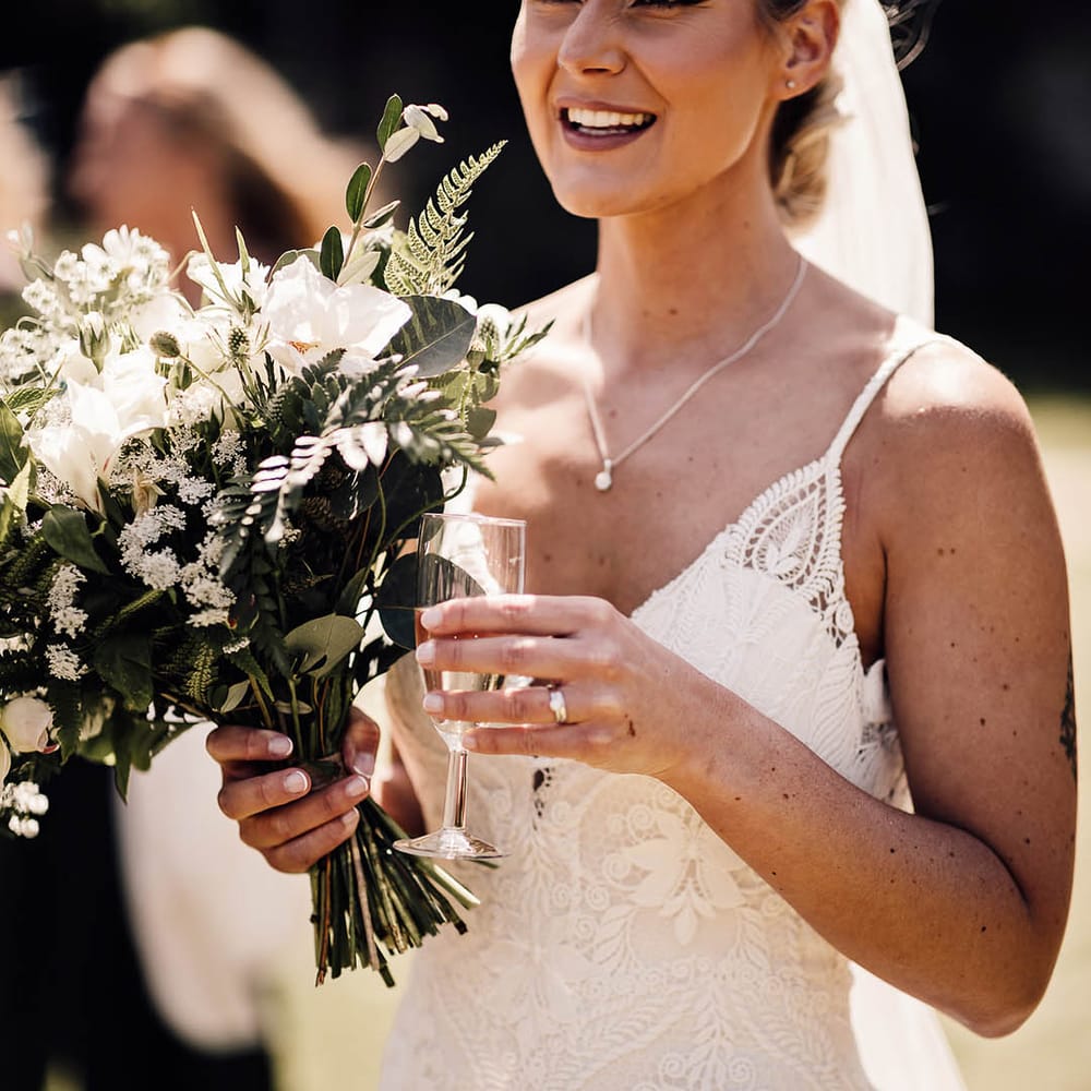 Bride with braided updo wearing lace wedding dress holding small glass of champagne and white flower bridal bouquet 