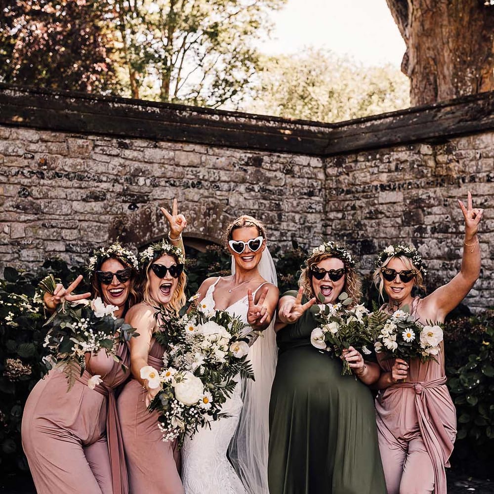 Bridesmaids and bride in retro heart sunglasses throw up peace signs with bridesmaids in pink and green dresses 